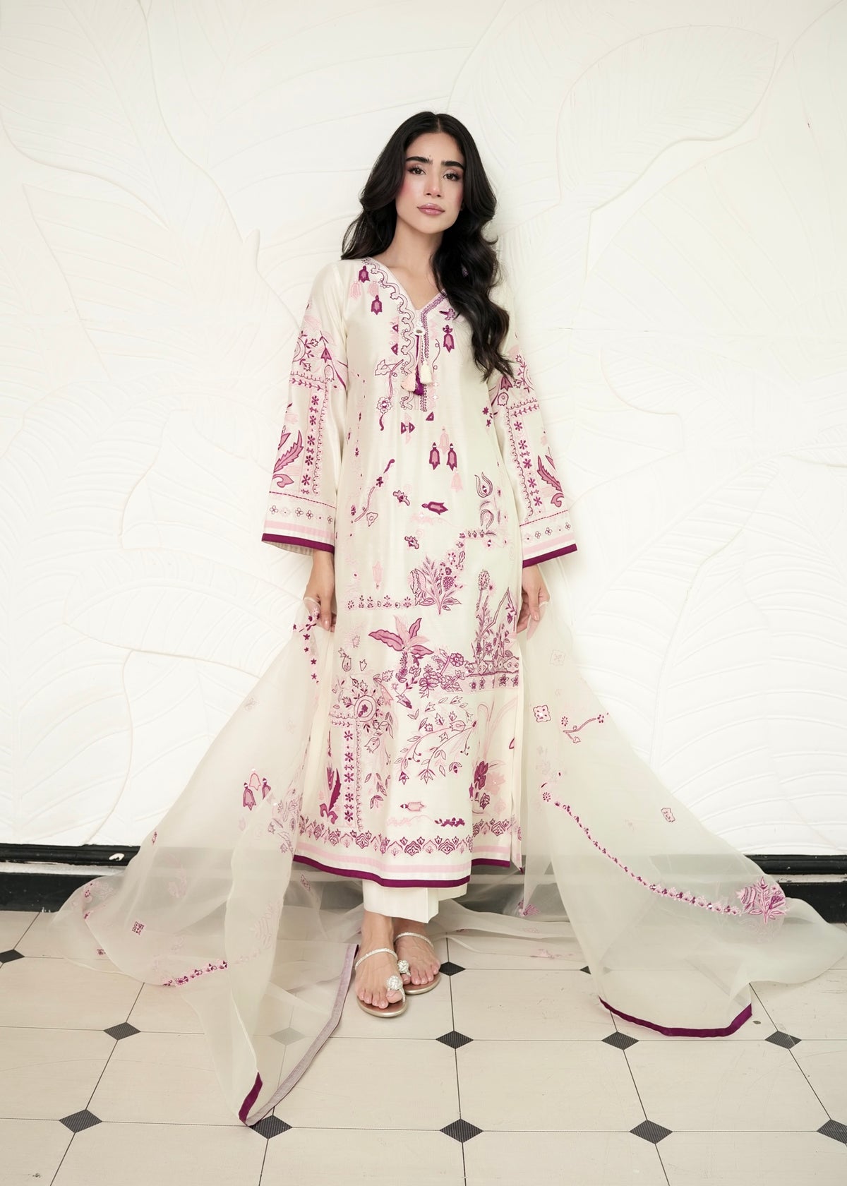 A woman modeling a traditional three-piece salwar kameez suit in off-white with pink floral embroidery, featuring a matching sheer dupatta, set against a textured white wall and a black-and-white checkered floor.