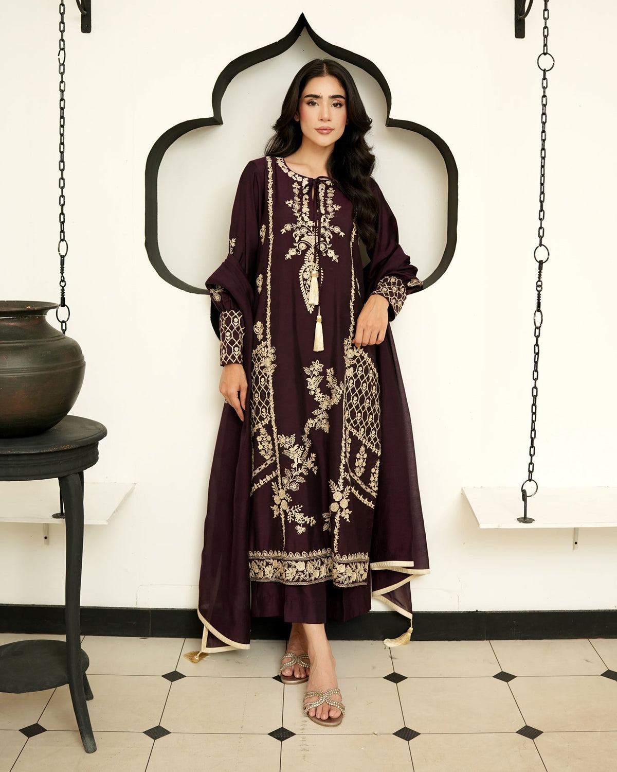 A woman modeling a long, dark plum embroidered kurta with beige floral patterns, standing in front of a white wall with a black arched frame.