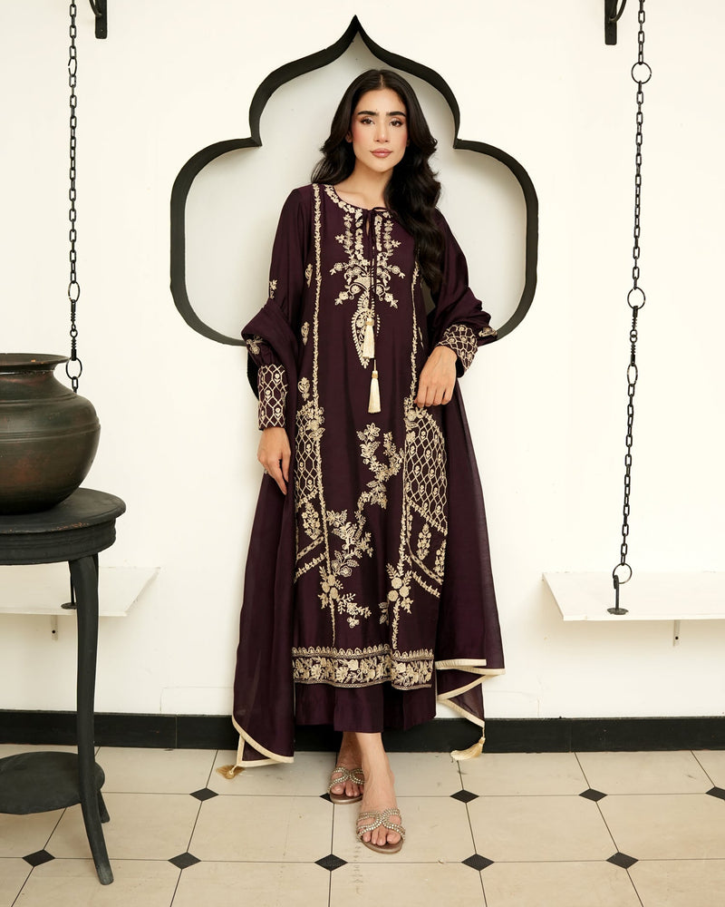 A woman modeling a long, dark plum embroidered kurta with beige floral patterns, standing in front of a white wall with a black arched frame.