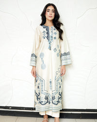 A woman modeling a cream-colored, long-sleeved embroidered tunic with blue patterns, standing against a white textured wall.