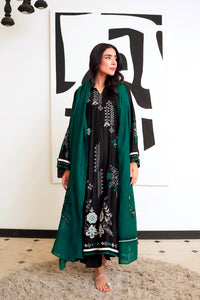 A woman stands indoors wearing a black embroidered long kurta with a deep green dupatta draped over her shoulders. The outfit has white and teal floral embroidery, and she is barefoot on a textured rug in front of a white wall with a large black abstract print.